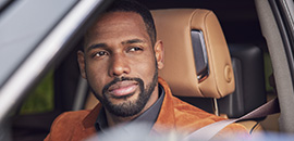 Man from the image above is sitting in the driver seat of a Cadillac and looking wistfully out the window.